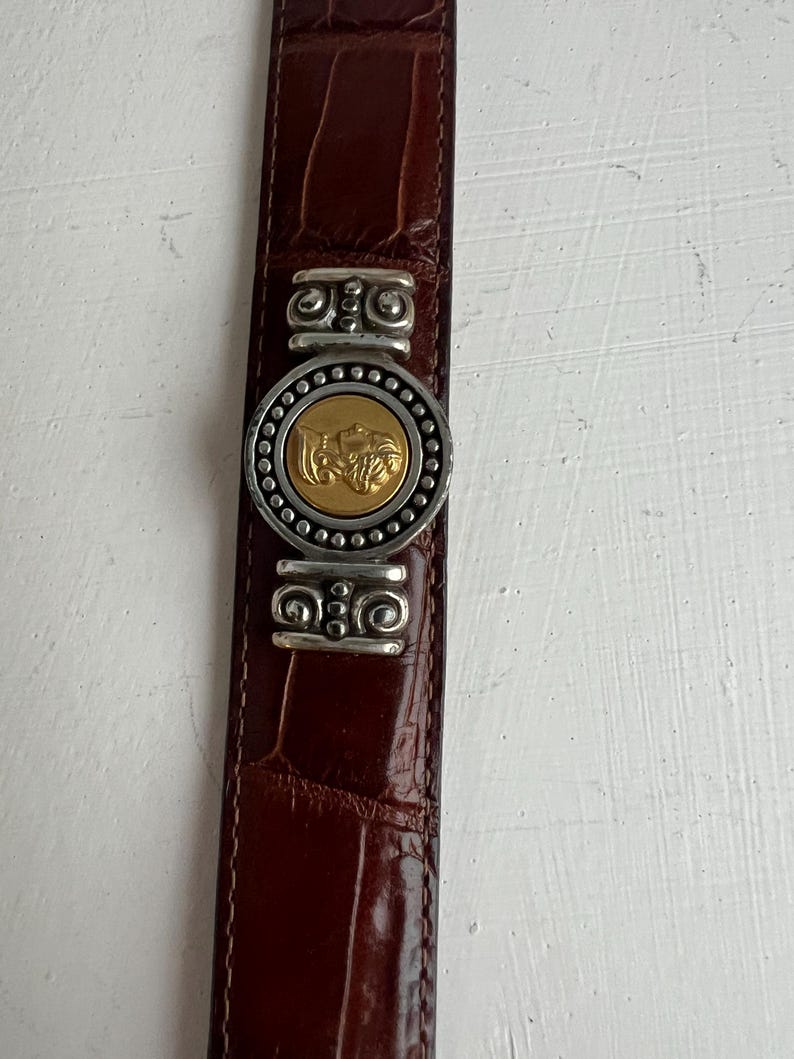 Puede incluir: Cintur&oacute;n de cuero marr&oacute;n con textura de cocodrilo. El cintur&oacute;n presenta una hebilla plateada con un medall&oacute;n dorado en el centro. La hebilla est&aacute; adornada con detalles decorativos plateados. Aproximadamente 3 cm de ancho.