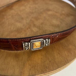 Puede incluir: Cintur&oacute;n de cuero marr&oacute;n con textura de cocodrilo. El cintur&oacute;n presenta una hebilla plateada con un cuadrado dorado en el centro. El cintur&oacute;n se muestra sobre una superficie de madera.