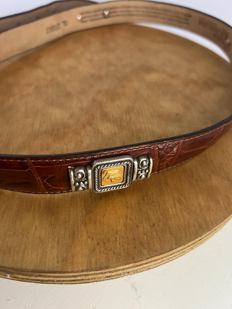 Puede incluir: Cintur&oacute;n de cuero marr&oacute;n con textura de cocodrilo. El cintur&oacute;n presenta una hebilla plateada con un cuadrado dorado en el centro. El cintur&oacute;n se muestra sobre una superficie de madera.