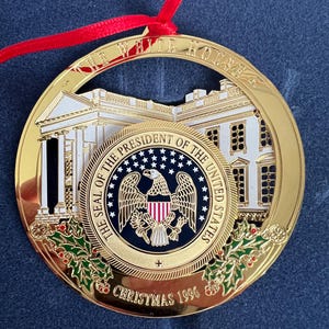 May include: A gold-colored Christmas ornament with a red ribbon. The ornament features a detailed depiction of the White House and the Seal of the President of the United States. The text "THE WHITE HOUSE" and "CHRISTMAS 1996" are visible.