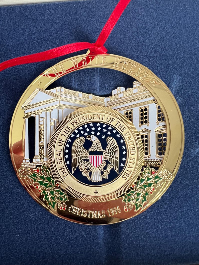 May include: A gold-colored Christmas ornament with a red ribbon. The ornament features a detailed depiction of the White House and the Seal of the President of the United States. The text "THE WHITE HOUSE" and "CHRISTMAS 1996" are visible.