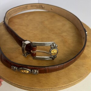 Puede incluir: Un cintur&oacute;n de cuero marr&oacute;n con hebilla plateada y detalles dorados. El cintur&oacute;n tiene una textura de piel de cocodrilo y est&aacute; enrollado sobre una superficie de madera. La hebilla presenta un emblema dorado.