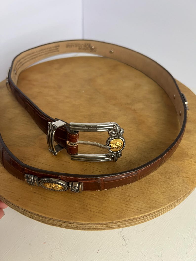 Puede incluir: Un cintur&oacute;n de cuero marr&oacute;n con hebilla plateada y detalles dorados. El cintur&oacute;n tiene una textura de piel de cocodrilo y est&aacute; enrollado sobre una superficie de madera. La hebilla presenta un emblema dorado.