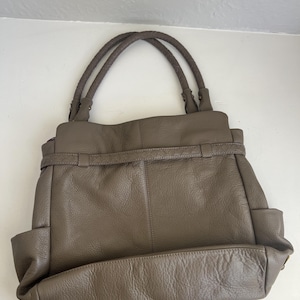 May include: A taupe leather handbag with braided handles. The bag has a structured design with a rectangular base, side pockets, and a belt detail across the front. The leather appears to have a slightly textured finish.