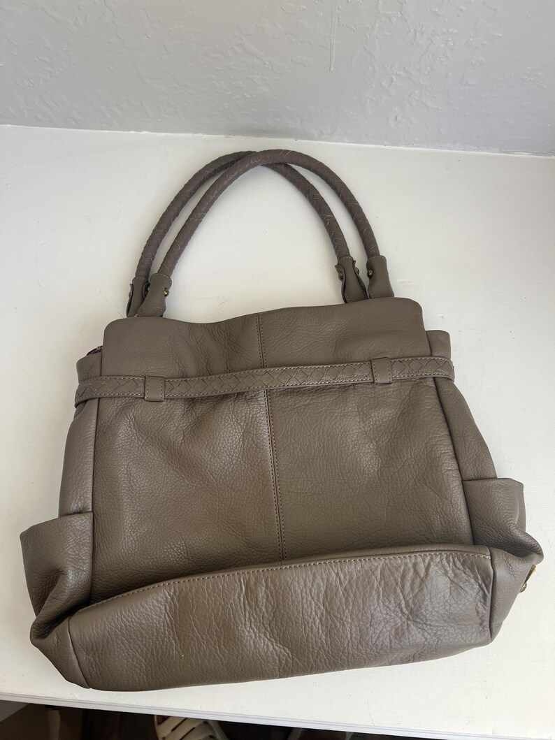 May include: A taupe leather handbag with braided handles. The bag has a structured design with a rectangular base, side pockets, and a belt detail across the front. The leather appears to have a slightly textured finish.