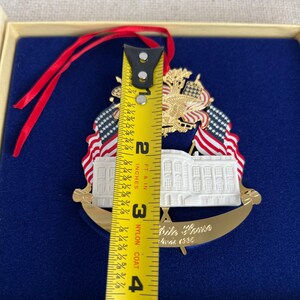 May include: A gold-colored White House ornament with red, white, and blue accents. The ornament features the White House, American flags, and an eagle. The ornament is approximately 3 inches (7.6 cm) tall.