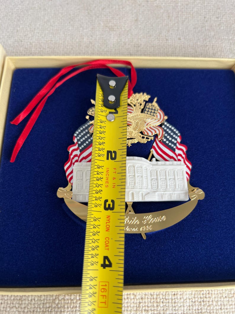 May include: A gold-colored White House ornament with red, white, and blue accents. The ornament features the White House, American flags, and an eagle. The ornament is approximately 3 inches (7.6 cm) tall.