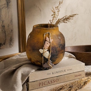 May include: A rustic, round, brown vase with a textured finish, holding dried wheat stalks. The vase is adorned with a decorative string and small, attached paper-like elements. It rests on a stack of books, including "THE KINFOLK HOME" and "PICASSO."