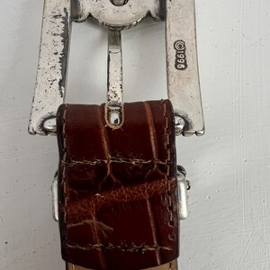 Puede incluir: Primer plano de un cintur&oacute;n de cuero marr&oacute;n con una hebilla plateada. La hebilla tiene un dise&ntilde;o decorativo y el a&ntilde;o 1996 est&aacute; estampado en el lateral. El cuero tiene un patr&oacute;n texturizado similar al de un cocodrilo.