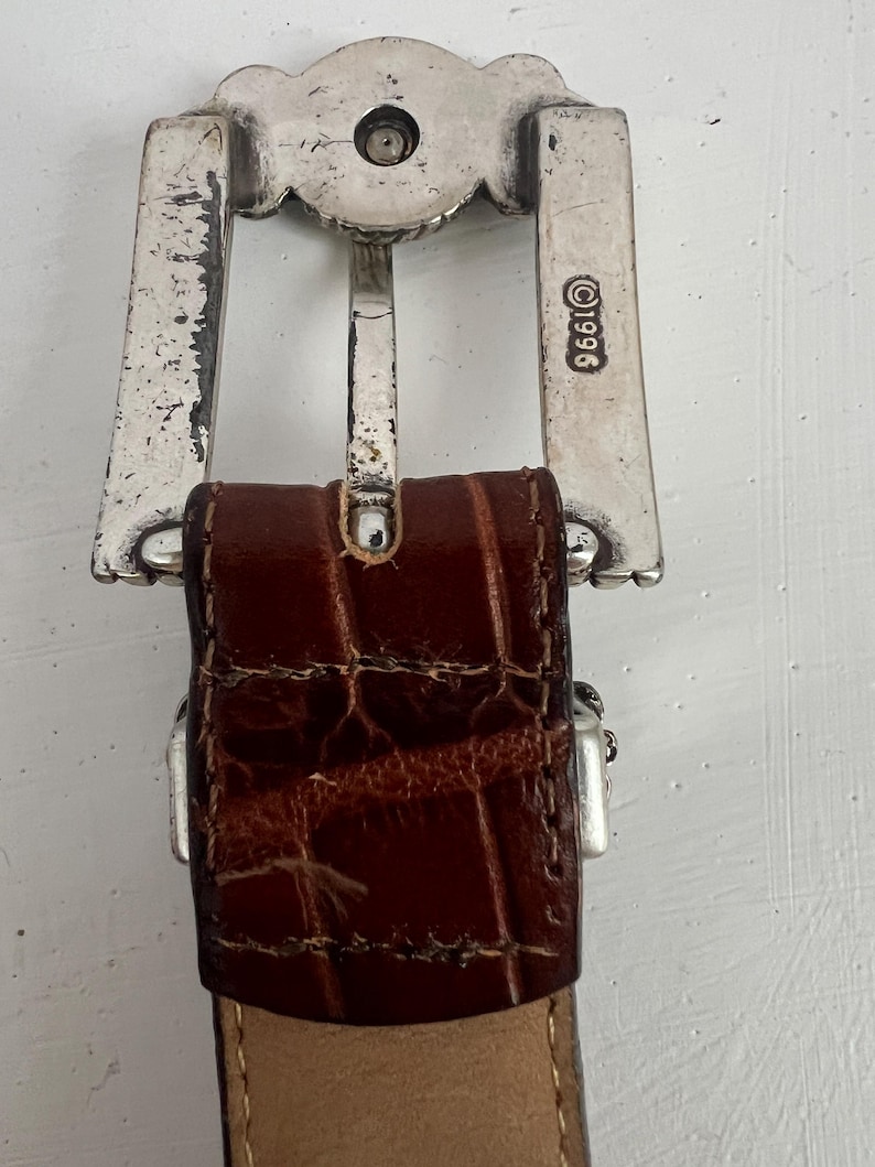 Puede incluir: Primer plano de un cintur&oacute;n de cuero marr&oacute;n con una hebilla plateada. La hebilla tiene un dise&ntilde;o decorativo y el a&ntilde;o 1996 est&aacute; estampado en el lateral. El cuero tiene un patr&oacute;n texturizado similar al de un cocodrilo.