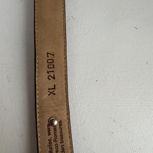 Puede incluir: Primer plano de un cintur&oacute;n de cuero marr&oacute;n con hebilla plateada. El interior del cintur&oacute;n es de color beige con el texto "XL 21007" y otro texto. El cintur&oacute;n es probablemente para pantalones.