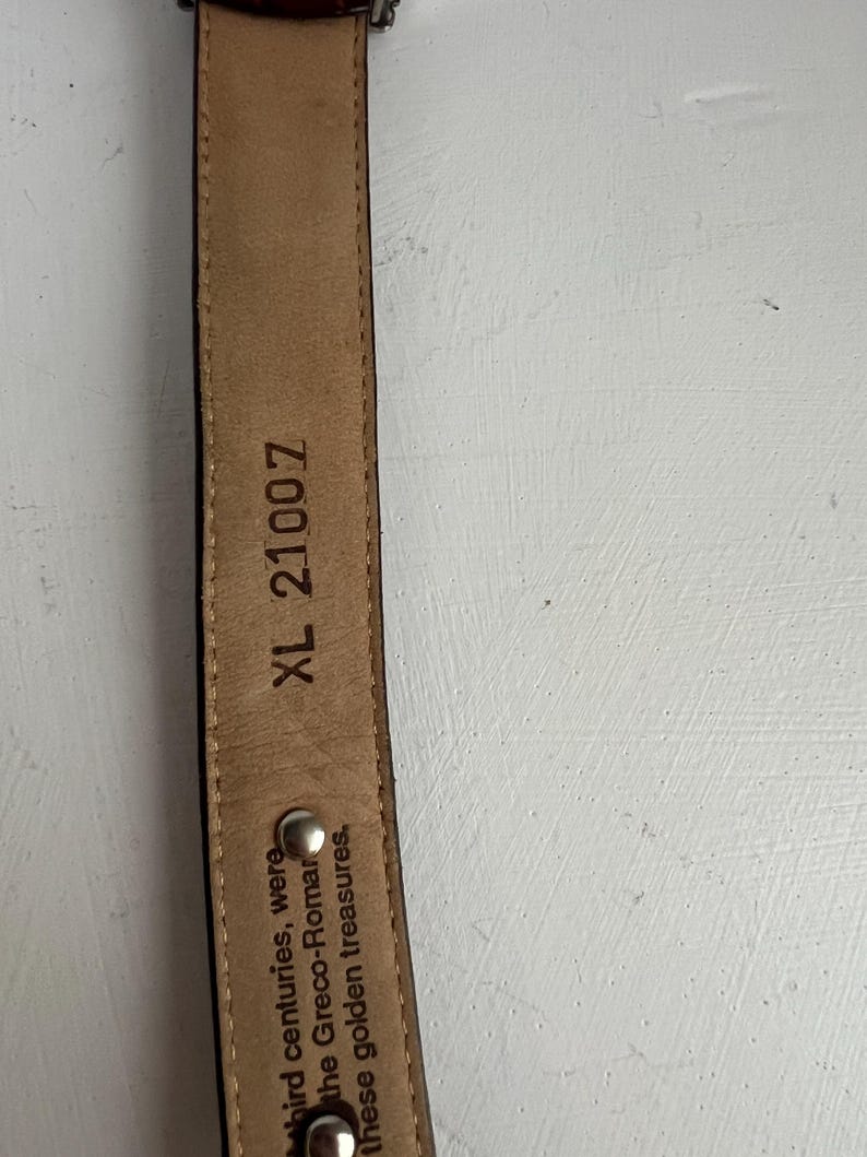 Puede incluir: Primer plano de un cintur&oacute;n de cuero marr&oacute;n con hebilla plateada. El interior del cintur&oacute;n es de color beige con el texto "XL 21007" y otro texto. El cintur&oacute;n es probablemente para pantalones.