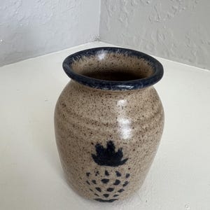 May include: A speckled beige ceramic vase with a dark blue rim and a dark blue pineapple design. The vase has a rounded body and a slightly flared opening. The design is centered on the front of the vase.