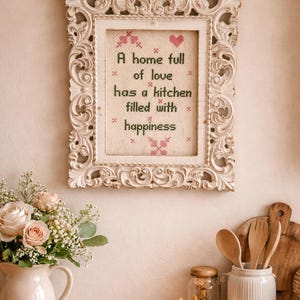 May include: A framed cross-stitch artwork with the quote "A home full of love has a kitchen filled with happiness." The frame is ornate and white. A bouquet of roses in a white pitcher and kitchen utensils are also visible.