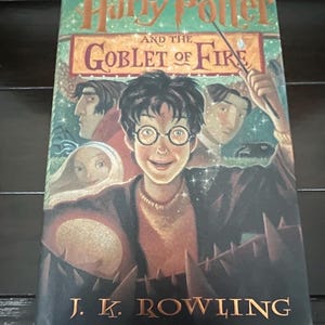 Puede incluir: Portada del libro "Harry Potter y el cáliz de fuego" de J.K. Rowling. La ilustración muestra a Harry Potter con gafas y otros personajes. El título es naranja y el nombre del autor dorado. Novela fantástica.