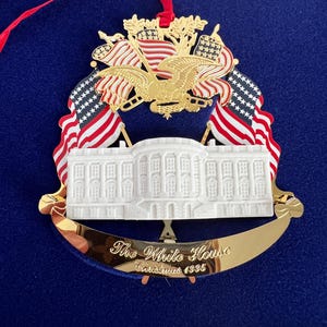 May include: A gold-toned Christmas ornament featuring the White House, American flags, and an eagle. The ornament has a red ribbon for hanging and the text "The White House Christmas 1995". The ornament is displayed on a blue background.