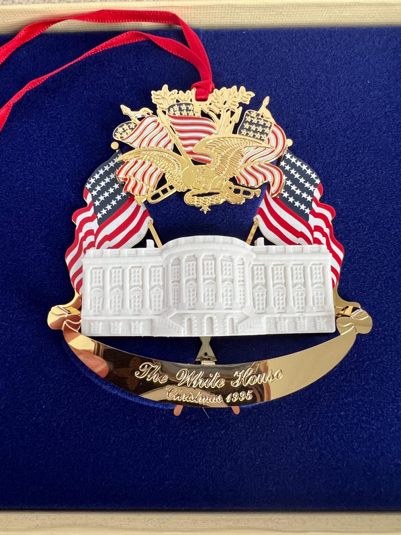 May include: A gold-toned Christmas ornament featuring the White House, American flags, and an eagle. The ornament has a red ribbon for hanging and the text "The White House Christmas 1995". The ornament is displayed on a blue background.