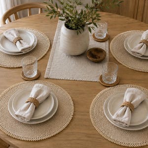 May include: A round wooden dining table set for four with beige woven placemats, white ceramic plates, light beige linen napkins held by braided rattan napkin rings, clear textured glass tumblers on small woven coasters, and a central ceramic vase.