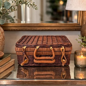 May include: A brown wicker picnic basket with a handle and clasp, next to a vase with greenery, books, and a candle. The basket is rectangular with a woven design and leather accents. The scene is set on a reflective surface.
