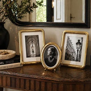 May include: A collection of gold-framed picture frames on a wooden table. The frames hold black and white photographs. A black vase with greenery and a marble tray are also on the table, along with books.