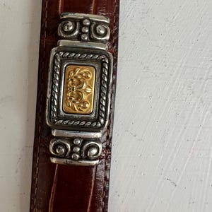 Puede incluir: Primer plano de un cintur&oacute;n de cuero marr&oacute;n con una hebilla decorativa plateada y dorada. La hebilla presenta un centro rectangular dorado con un dise&ntilde;o ornamentado, enmarcado por detalles plateados. El cintur&oacute;n tiene un patr&oacute;n texturizado similar al de un cocodrilo.