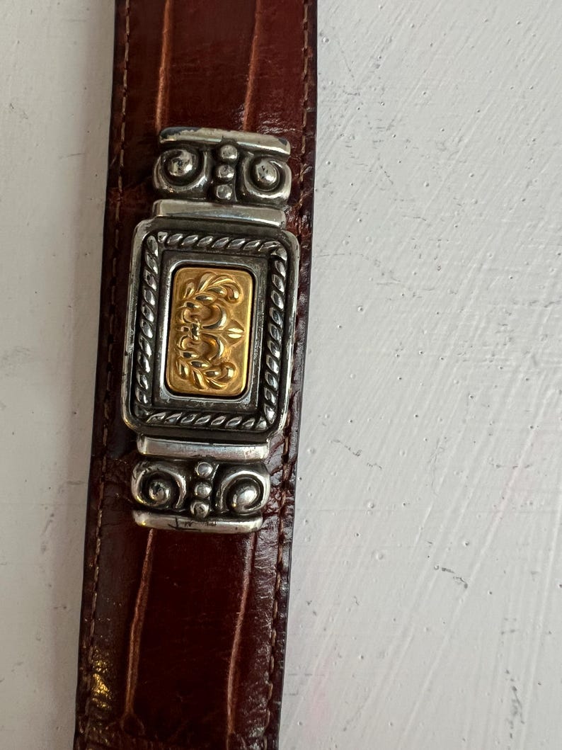 Puede incluir: Primer plano de un cintur&oacute;n de cuero marr&oacute;n con una hebilla decorativa plateada y dorada. La hebilla presenta un centro rectangular dorado con un dise&ntilde;o ornamentado, enmarcado por detalles plateados. El cintur&oacute;n tiene un patr&oacute;n texturizado similar al de un cocodrilo.