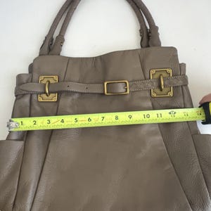 May include: Taupe leather handbag with double handles and gold-tone hardware. The bag features a buckle closure and side pockets. The width of the bag is approximately 13 inches.