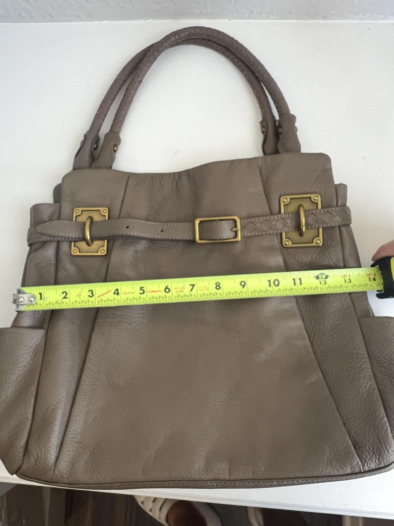 May include: Taupe leather handbag with double handles and gold-tone hardware. The bag features a buckle closure and side pockets. The width of the bag is approximately 13 inches.