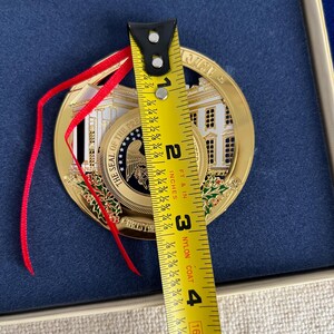 May include: A gold-colored Christmas ornament featuring the White House and the words "THE SEAL OF THE PRESIDENT" and "CHRISTMAS". The ornament is approximately 3 inches in diameter and has a red ribbon for hanging.