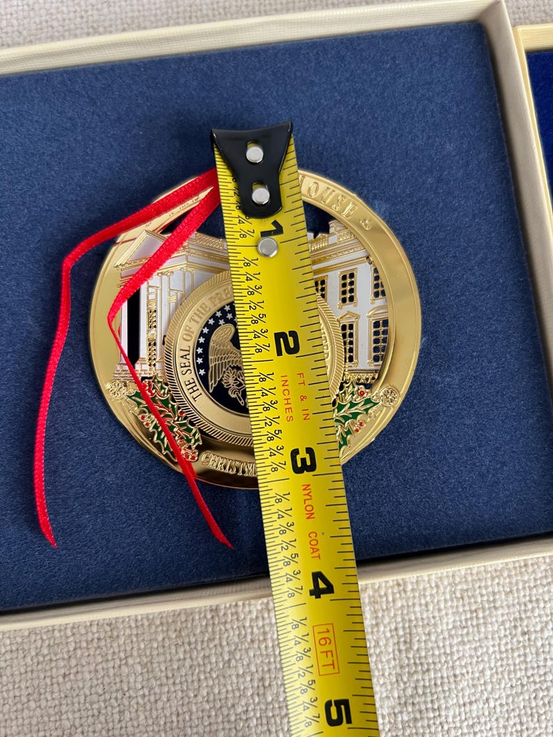 May include: A gold-colored Christmas ornament featuring the White House and the words "THE SEAL OF THE PRESIDENT" and "CHRISTMAS". The ornament is approximately 3 inches in diameter and has a red ribbon for hanging.