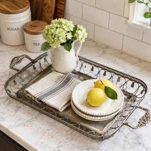 May include: A rectangular metal serving tray with ornate handles and a lattice-style rim. Inside the tray sits a white ceramic pitcher filled with green hydrangea flowers, a folded beige linen napkin with gray stripes, and two white plates holding two yellow lemons.