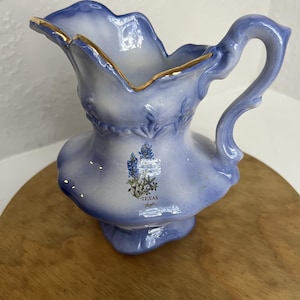 Könnte beinhalten: Hellblaue Keramikkrug mit gerüschtem Rand und gebogenem Griff. Der Krug hat einen goldenen Rand und ein Blumendesign mit dem Wort "TEXAS" auf der Vorderseite. Der Krug steht auf einer Holzoberfläche.