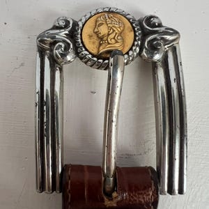 Puede incluir: Hebilla de cintur&oacute;n de metal plateado con un medall&oacute;n dorado que presenta el perfil de una persona. La hebilla tiene un dise&ntilde;o decorativo y est&aacute; sujeta a un cintur&oacute;n de cuero marr&oacute;n. La hebilla mide aproximadamente 7,6 cm de ancho.