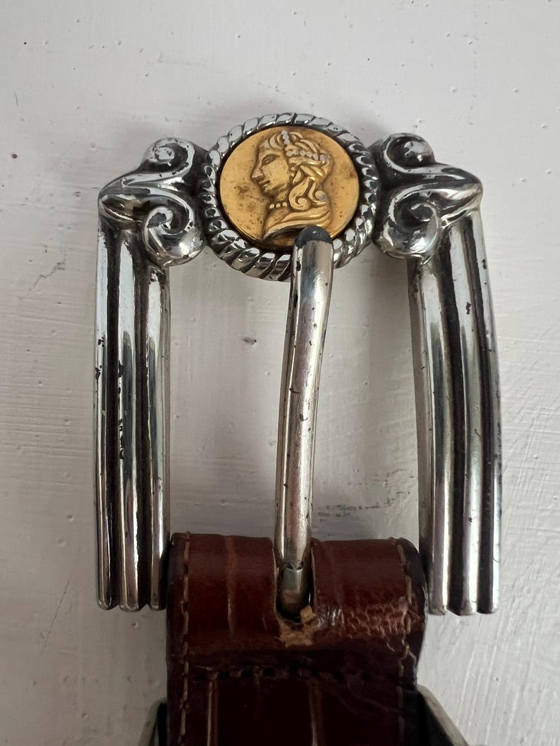 Puede incluir: Hebilla de cintur&oacute;n de metal plateado con un medall&oacute;n dorado que presenta el perfil de una persona. La hebilla tiene un dise&ntilde;o decorativo y est&aacute; sujeta a un cintur&oacute;n de cuero marr&oacute;n. La hebilla mide aproximadamente 7,6 cm de ancho.