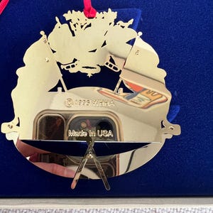 May include: A gold-colored, decorative ornament with a cut-out design featuring flags and a sports theme. The ornament has a red ribbon for hanging and the text "Made in USA". The year "1995" is also visible.