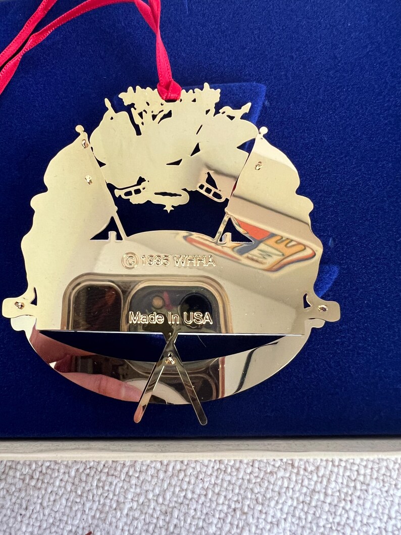 May include: A gold-colored, decorative ornament with a cut-out design featuring flags and a sports theme. The ornament has a red ribbon for hanging and the text "Made in USA". The year "1995" is also visible.