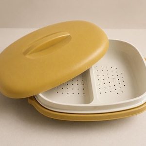 Puede incluir: Contenedor de alimentos de plástico amarillo mostaza y blanco con tapa. La tapa es de forma ovalada con asa. El interior es blanco con sección perforada. El contenedor está abierto.
