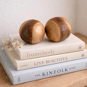 Può includere: Due sfere decorative in legno con fasce marroni chiare e scure alternate poggiano su una pila di libri. I libri hanno titoli come "homebody" e "Live Beautiful". L'estetica generale è minimalista e naturale.