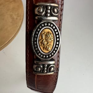 Puede incluir: Cintur&oacute;n de cuero marr&oacute;n con una hebilla decorativa plateada y dorada. La hebilla presenta una pieza central ovalada dorada con un dise&ntilde;o floral, rodeada por un marco plateado. El cintur&oacute;n tiene una textura de piel de cocodrilo.