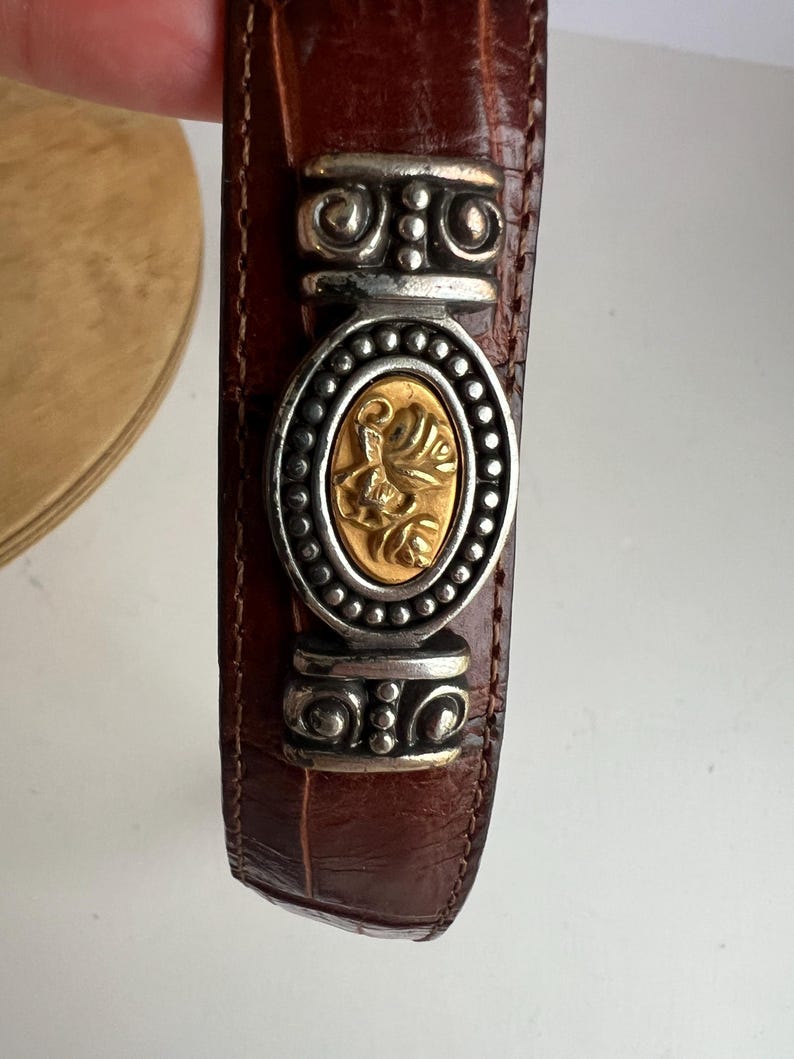 Puede incluir: Cintur&oacute;n de cuero marr&oacute;n con una hebilla decorativa plateada y dorada. La hebilla presenta una pieza central ovalada dorada con un dise&ntilde;o floral, rodeada por un marco plateado. El cintur&oacute;n tiene una textura de piel de cocodrilo.