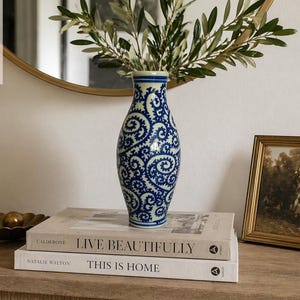 May include: A blue and white patterned vase holds olive branches, resting on a stack of books titled "Live Beautifully" and "This is Home." A gold-framed picture and a decorative bowl are also on the wooden surface, with a gold-rimmed mirror in the background.
