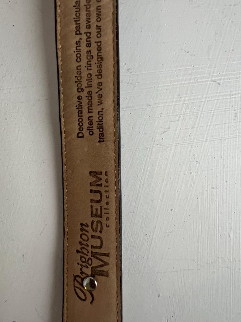 Puede incluir: Primer plano del interior de un cintur&oacute;n de cuero marr&oacute;n con el texto "Brighton MUSEUM collection". El interior del cintur&oacute;n tambi&eacute;n presenta el texto: "Monedas de oro decorativas, particularmente a menudo convertidas en anillos y premiadas por la tradici&oacute;n, hemos dise&ntilde;ado las nuestras."