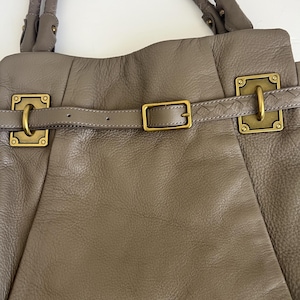 May include: Taupe leather handbag with a braided handle and gold-colored hardware. The bag features a belt closure with a buckle and decorative rectangular clasps. The leather has a textured appearance.