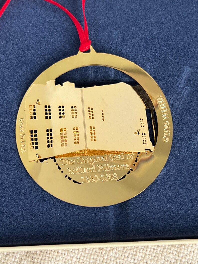 May include: A gold-colored Christmas ornament with a building silhouette and the text "The Original Seal of Millard Fillmore 1850-1853." The ornament is circular with a red ribbon for hanging.