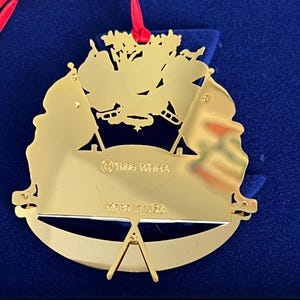 May include: A gold-colored Christmas ornament with a red ribbon. The ornament features a design with flags and the text "Made in USA" and "&copy; 1995 WHHA". The ornament is displayed on a blue surface.
