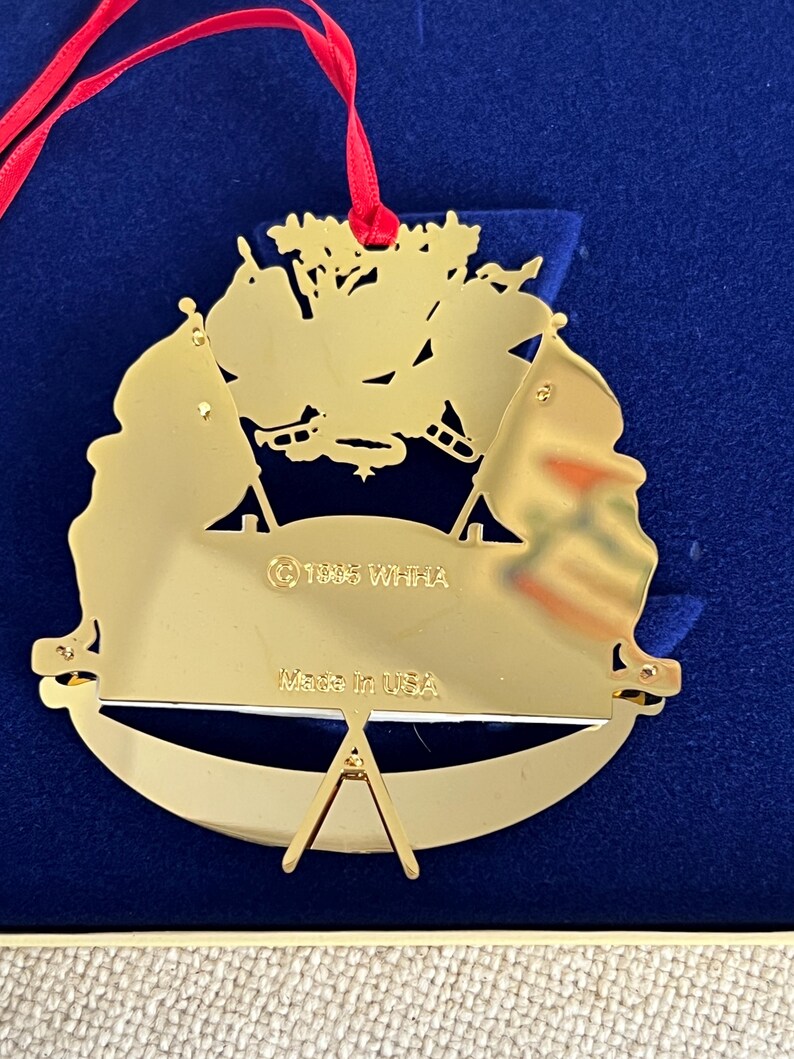 May include: A gold-colored Christmas ornament with a red ribbon. The ornament features a design with flags and the text "Made in USA" and "&copy; 1995 WHHA". The ornament is displayed on a blue surface.