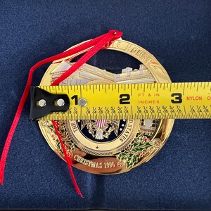 May include: A gold-colored Christmas ornament with a red ribbon. The ornament features an image of the White House and the text "THE WHITE HOUSE" and "CHRISTMAS 1996". The ornament is approximately 2.75 inches in diameter.