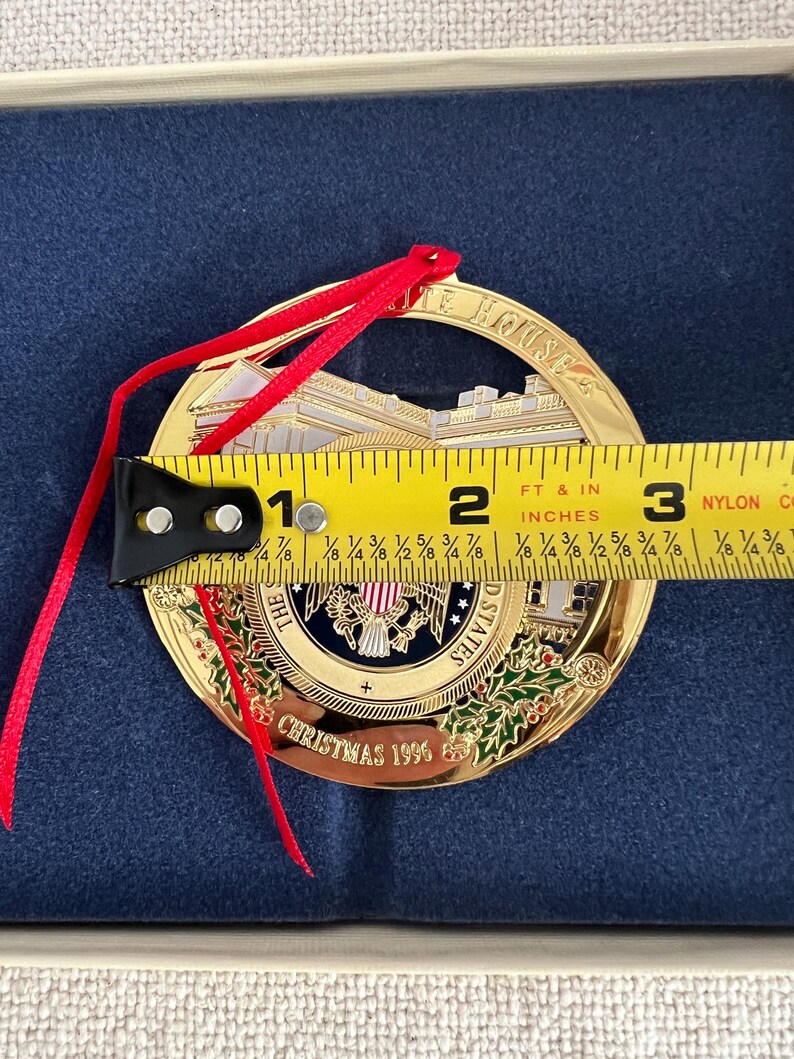 May include: A gold-colored Christmas ornament with a red ribbon. The ornament features an image of the White House and the text "THE WHITE HOUSE" and "CHRISTMAS 1996". The ornament is approximately 2.75 inches in diameter.