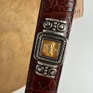 Puede incluir: Cintur&oacute;n de cuero marr&oacute;n con hebilla plateada y dorada. La hebilla presenta un dise&ntilde;o de corona dorada dentro de un marco plateado. El cintur&oacute;n tiene un patr&oacute;n texturizado similar al de un cocodrilo.