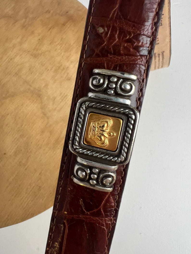 Puede incluir: Cintur&oacute;n de cuero marr&oacute;n con hebilla plateada y dorada. La hebilla presenta un dise&ntilde;o de corona dorada dentro de un marco plateado. El cintur&oacute;n tiene un patr&oacute;n texturizado similar al de un cocodrilo.